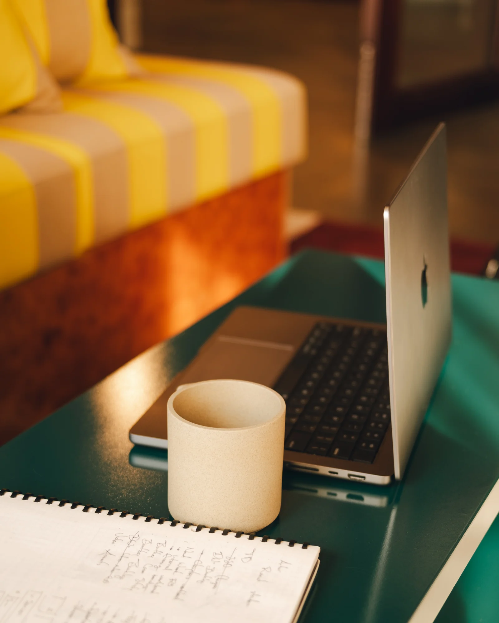 A laptop, a mug and a notebook of handwritten notes on a low table in the studio