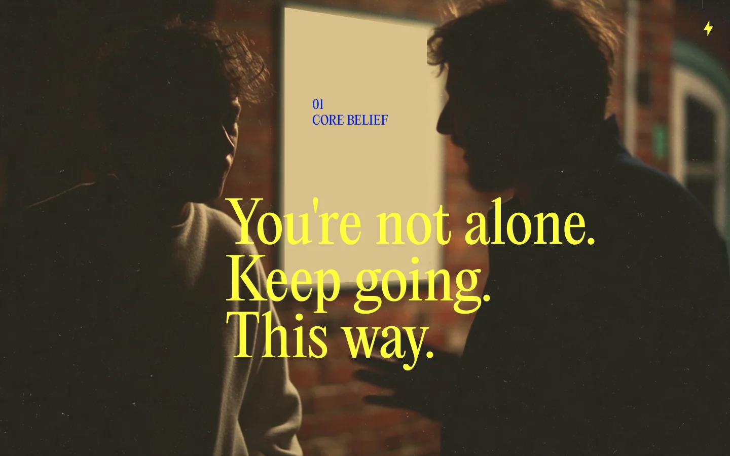 The core belief slide: You're not alone. Keep going. This way., set in yellow serif over two people talking in a warm-lit room